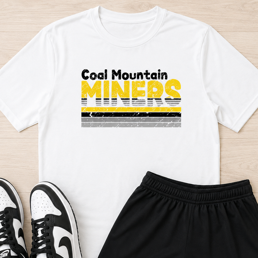 A flat-lay outfit consisting of a white t-shirt with a 'Coal Mountain MINERS' graphic, black athletic shorts, and black-and-white sneakers, arranged on a light wood floor.