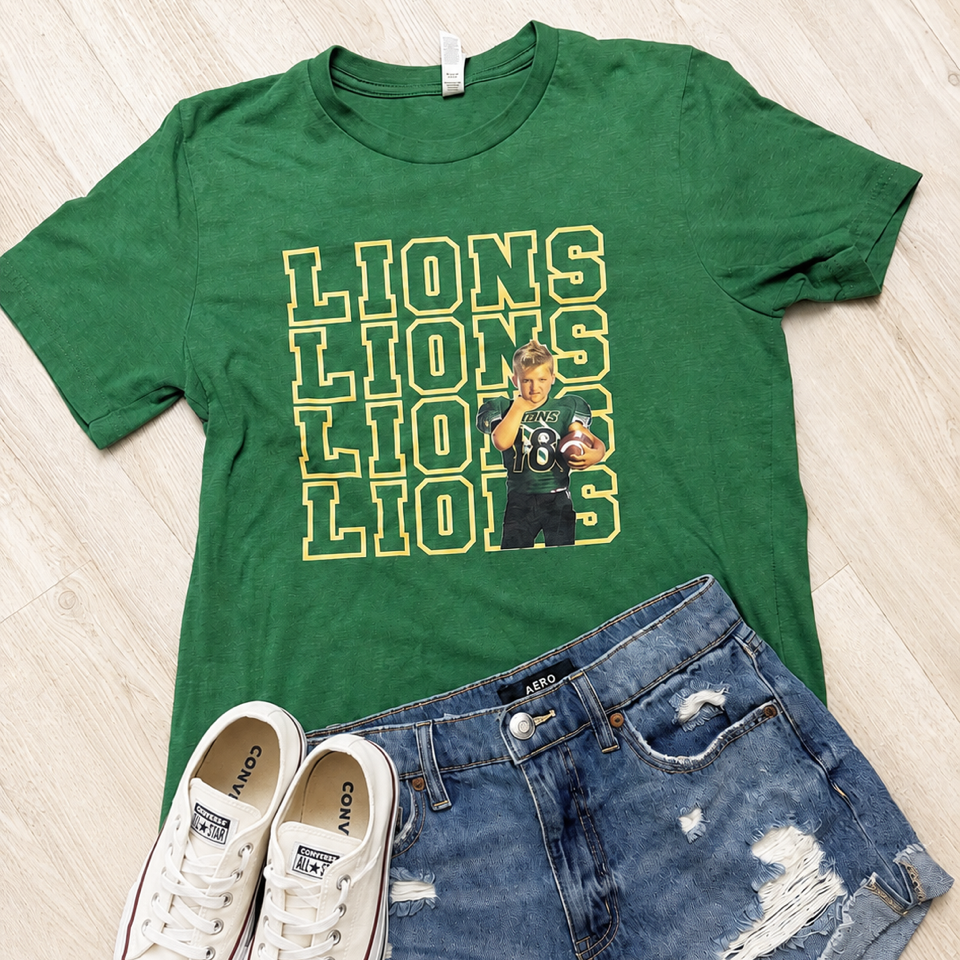 A green graphic t-shirt with 'LIONS' printed in yellow and white, styled in a flat lay with blue denim shorts and white canvas sneakers on a light wood floor.