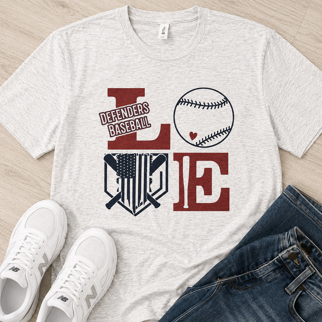 A heather light gray graphic T-shirt laid flat on a wood floor, styled with white sneakers and blue jeans. The shirt graphic reads "Defenders Baseball" and uses the letters L, O, and E with a baseball and shield design in dark red and navy.