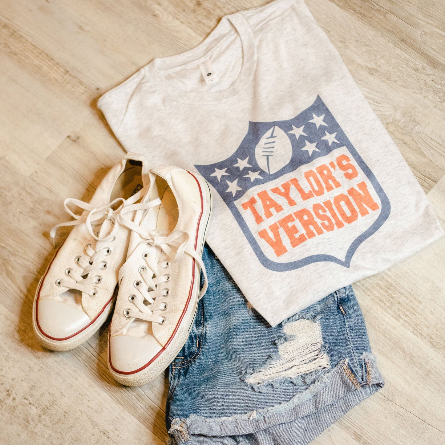 A heather white t-shirt with a graphic of stars and the text 'Taylor's Version' on the front, displayed alongside a pair of white sneakers and blue denim shorts with a frayed hem.