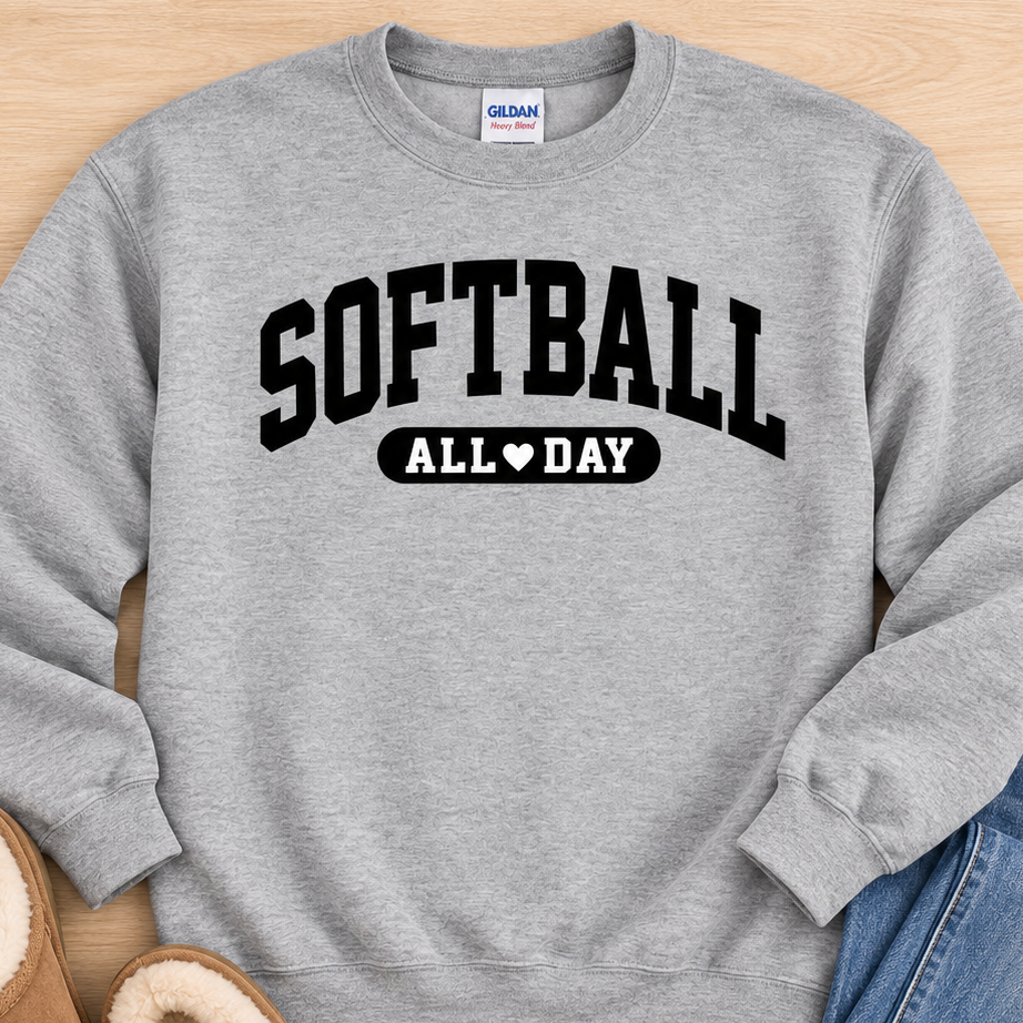 A heather gray crewneck sweatshirt with 'SOFTBALL ALL DAY' printed in black on the front, styled in a flat lay with blue jeans and tan slip-on slippers on a light wood background. The Gildan brand tag is visible inside the collar.