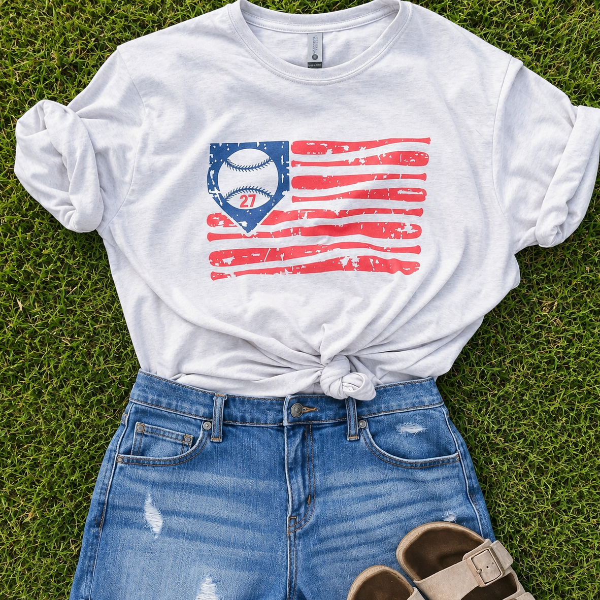 A white graphic t-shirt with a distressed red, white, and blue baseball flag design, styled in a flat lay on green grass with blue denim shorts and beige sandals.