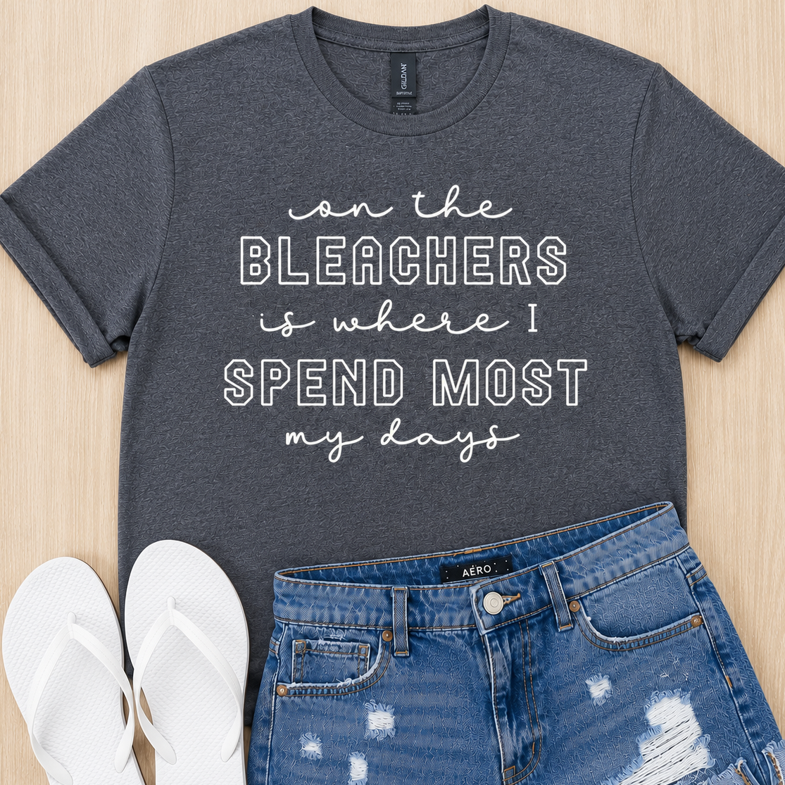 A dark heather gray graphic t-shirt with white text reading 'on the BLEACHERS is where I SPEND MOST my days', styled in a flat lay with blue distressed denim shorts and white flip-flops on a light wood background.