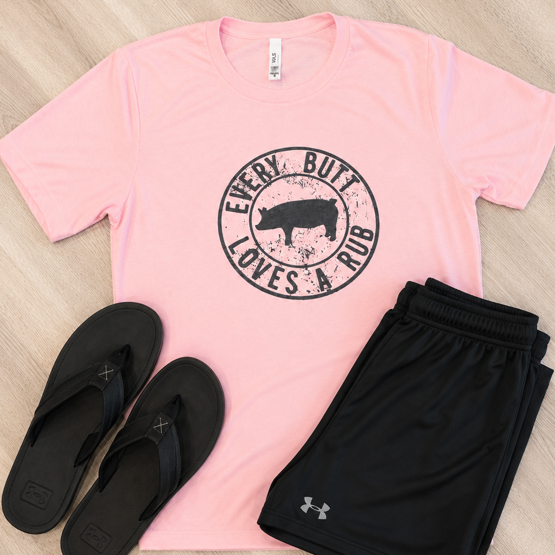A pink short-sleeve graphic T-shirt laid flat on a light wood floor, paired with black shorts and black flip-flops. The shirt has a circular distressed graphic with a pig silhouette and the words “EVERY BULL LOVES A HUG.
