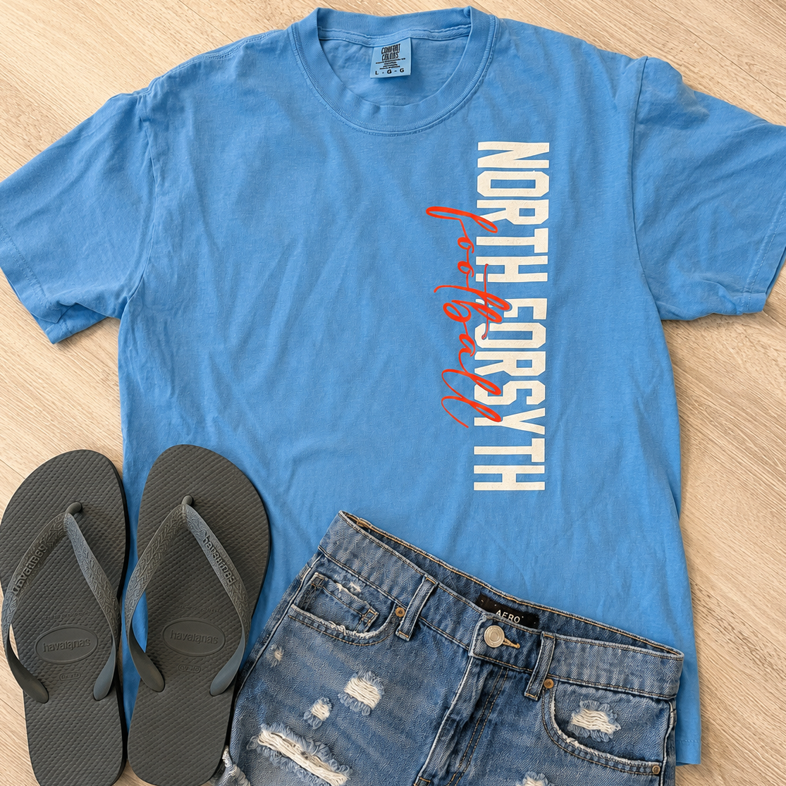 A flat-lay outfit featuring a light blue t-shirt with 'NORTH ENGLESYTH' printed vertically in white and red script, styled with distressed denim shorts and black flip-flops on a light wood floor.