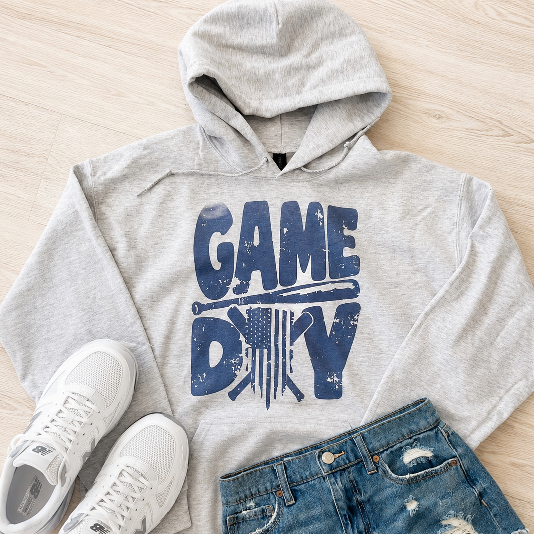 A styled flat-lay outfit featuring a light heather gray hoodie with a distressed blue 'GAME DAY' graphic, paired with blue distressed denim shorts and white sneakers on a light wood floor background.