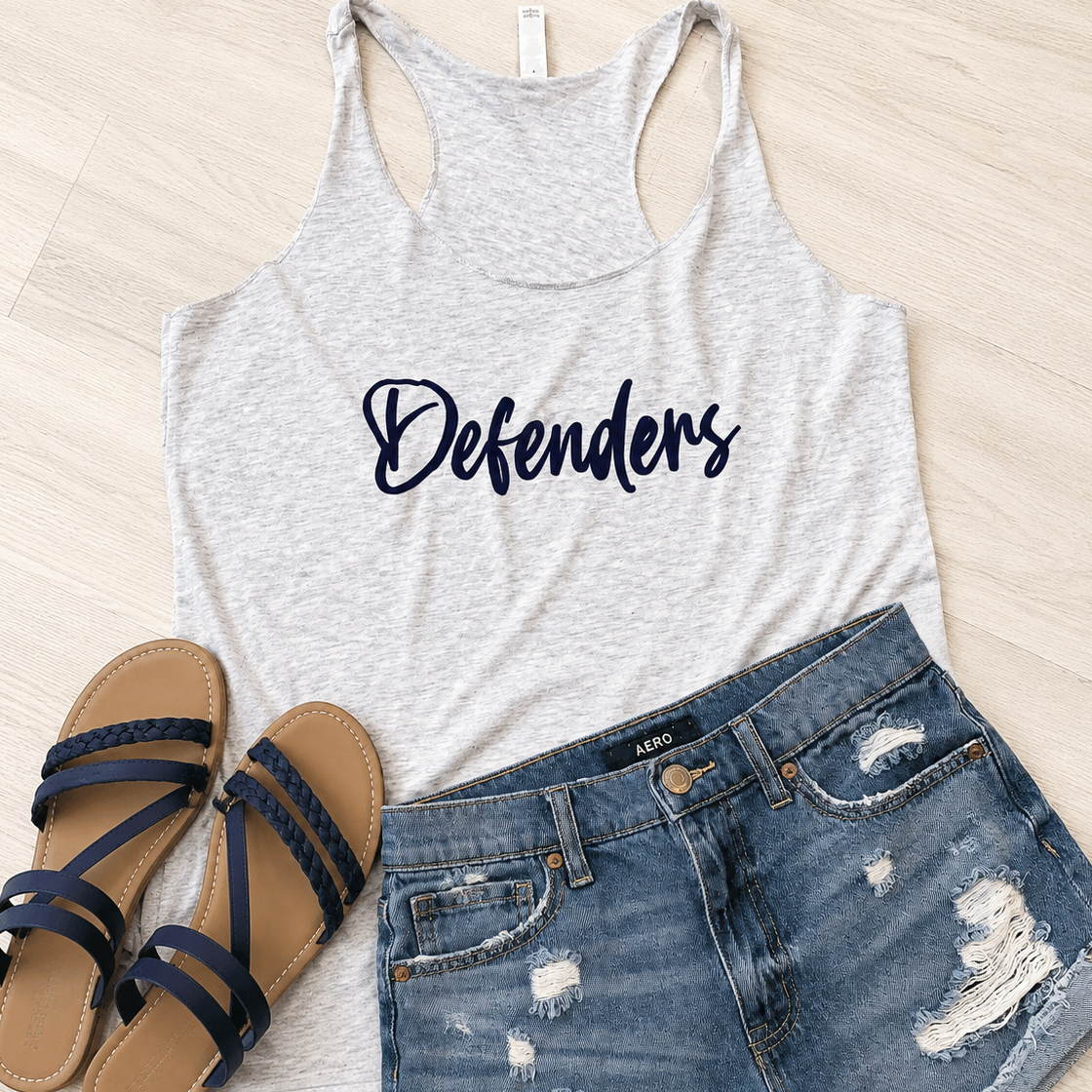 A light heather gray racerback tank top with the word 'Defenders' printed in navy script across the chest, styled in a flat lay with distressed blue denim shorts and navy strappy sandals on a light wood floor.