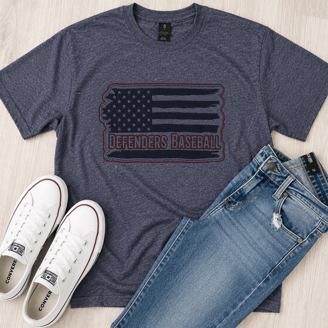 A heathered navy blue graphic t-shirt featuring a distressed American flag design with the text 'DEFENDERS BASEBALL' in red and navy. The t-shirt is styled in a flat lay on a light wood floor alongside a pair of blue jeans and white Converse sneakers.
