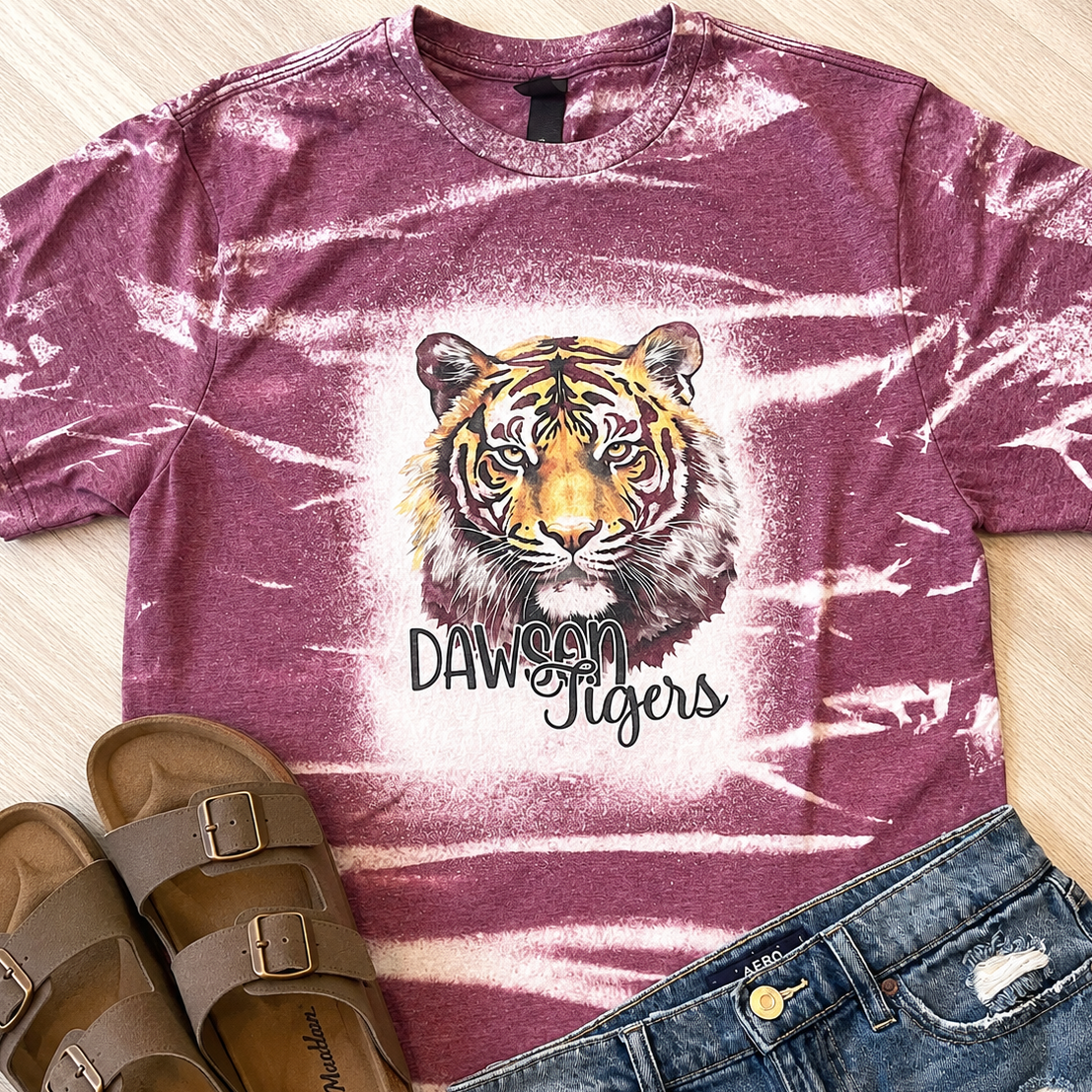 A mauve tie-dye t-shirt with a tiger graphic and the text 'DAWSON Tigers' laid flat on a light wood floor, styled with brown slide sandals and blue jeans.