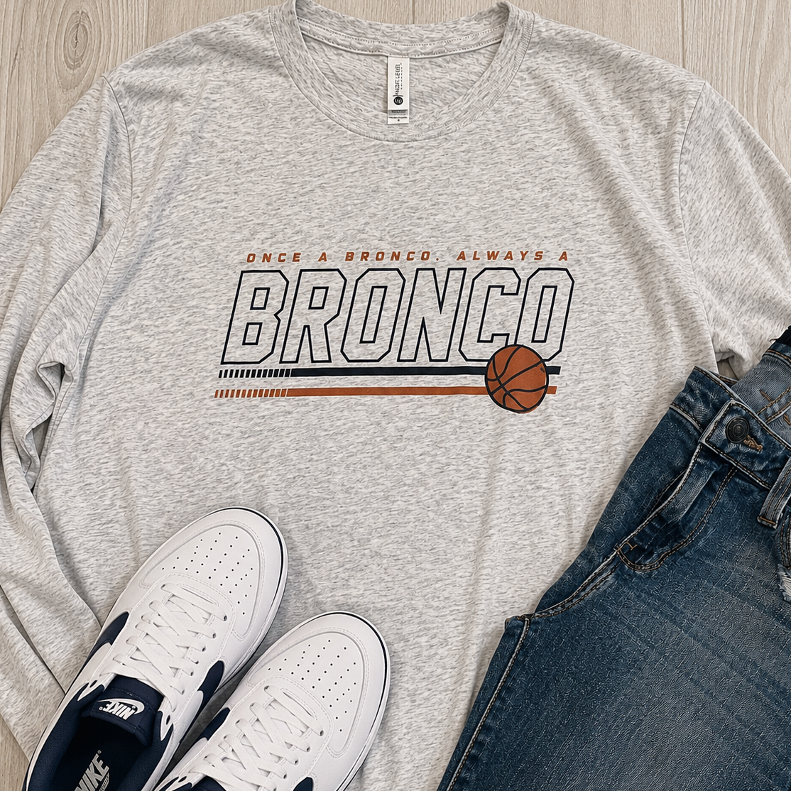 A heather gray long-sleeve graphic T-shirt laid flat on a light wood surface, styled with white Nike sneakers and blue jeans. The shirt has a large 'BRONCO' graphic with a basketball and small orange text above it.