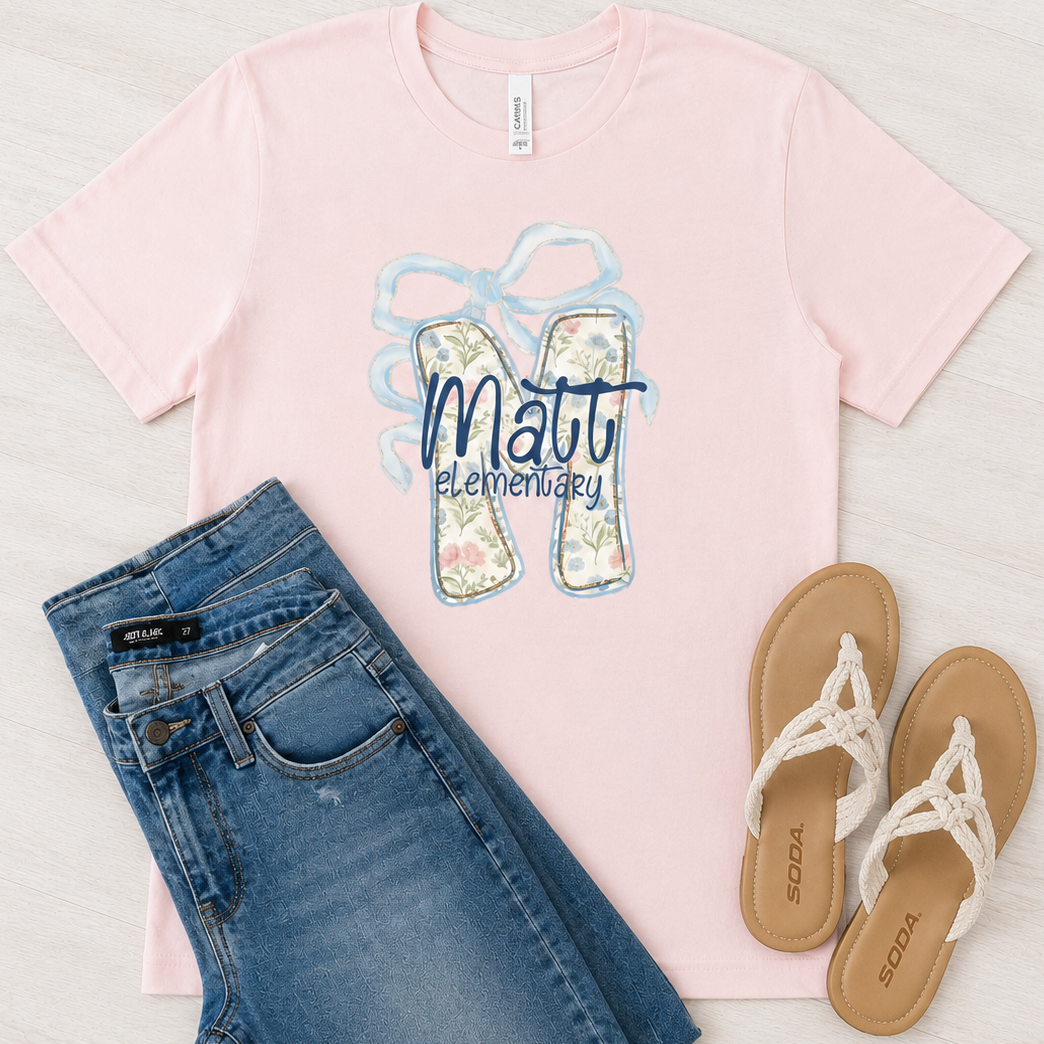 A flat-lay outfit arrangement featuring a light pink short-sleeve t-shirt with a 'Matt Elementary' flip-flop graphic, styled with blue jeans and tan sandals on a light wood background.