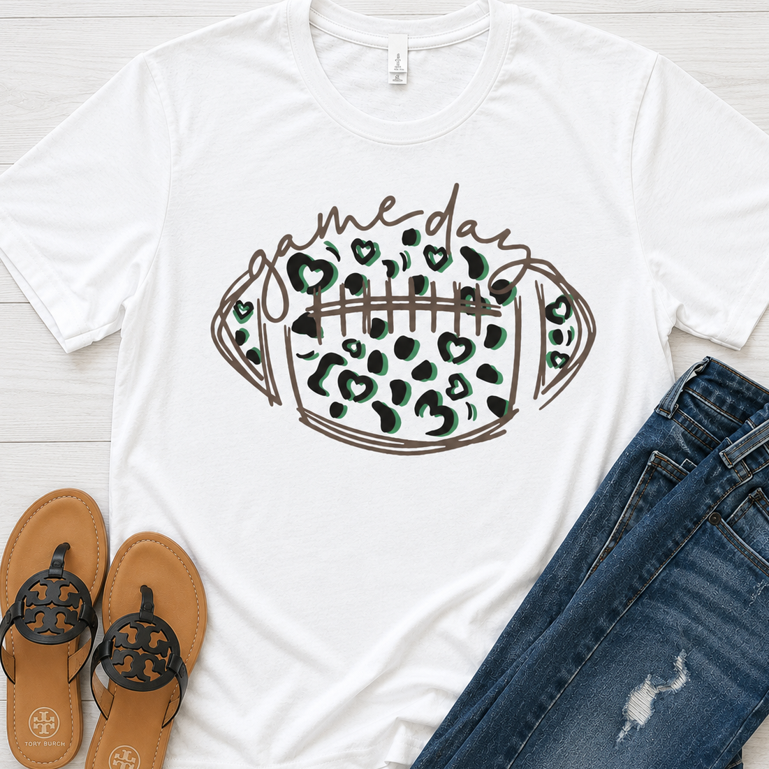 A white short-sleeve graphic T-shirt laid flat on a light wood surface, styled with blue jeans and tan sandals. The shirt features a football-shaped graphic with leopard print and the words 'game day' across the front.