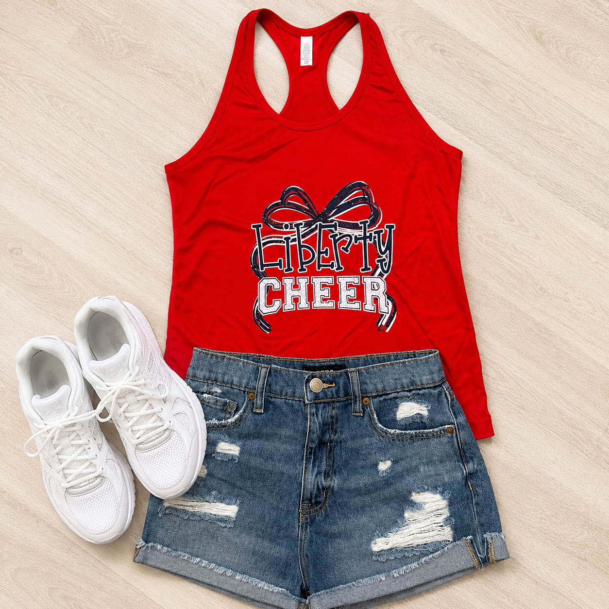 A red racerback tank top with a 'Liberty Cheer' ribbon graphic, styled in a flat lay with distressed blue denim shorts and white sneakers on a light wood background.