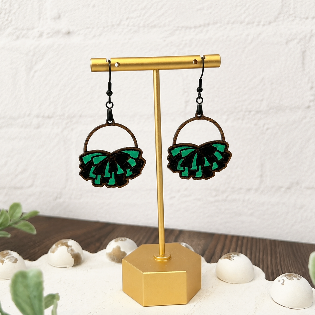 A pair of dangling hoop earrings displayed on a gold stand. The earrings have dark brown hoops with layered green and black ruffled petal-like charms, hanging against a white brick wall background with decorative plants and white stones.