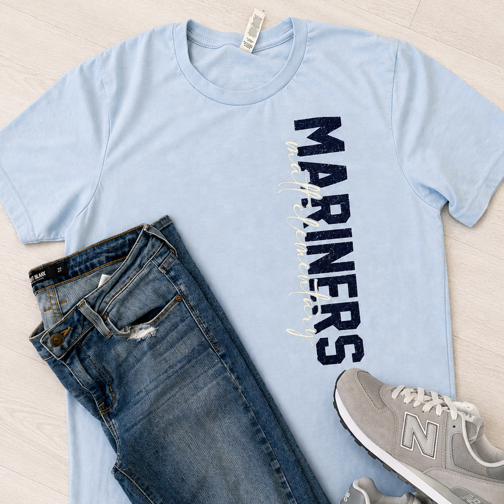 A light blue graphic T-shirt with 'MARINERS' printed vertically in dark blue, styled in a flat lay with distressed blue jeans and gray New Balance sneakers on a light wood floor.