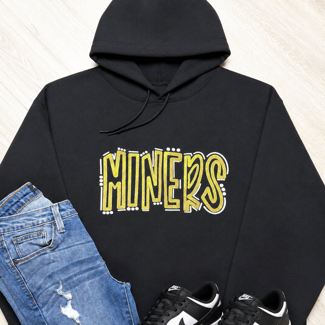 A black pullover hoodie with a yellow and white 'MINERS' graphic on the chest, styled in a flat lay with blue jeans and black-and-white Nike sneakers on a light wood background.