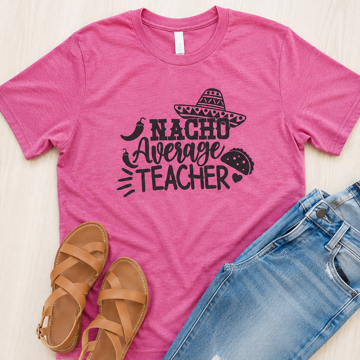 A pink short-sleeve graphic T-shirt laid flat on a light wood surface, styled with tan sandals and blue jeans. The shirt has a black graphic reading “NACHO Average TEACHER” with a sombrero, chili pepper, and taco illustration.