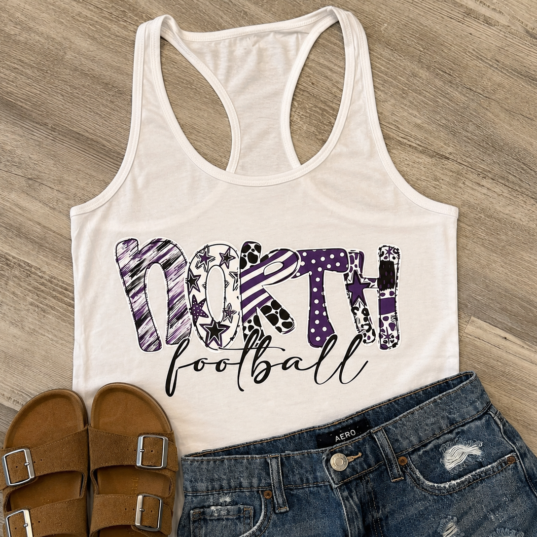 A white racerback tank top with a purple and black patterned 'NORTH football' graphic, styled in a flat lay with blue distressed denim shorts and brown slide sandals on a wood floor background.