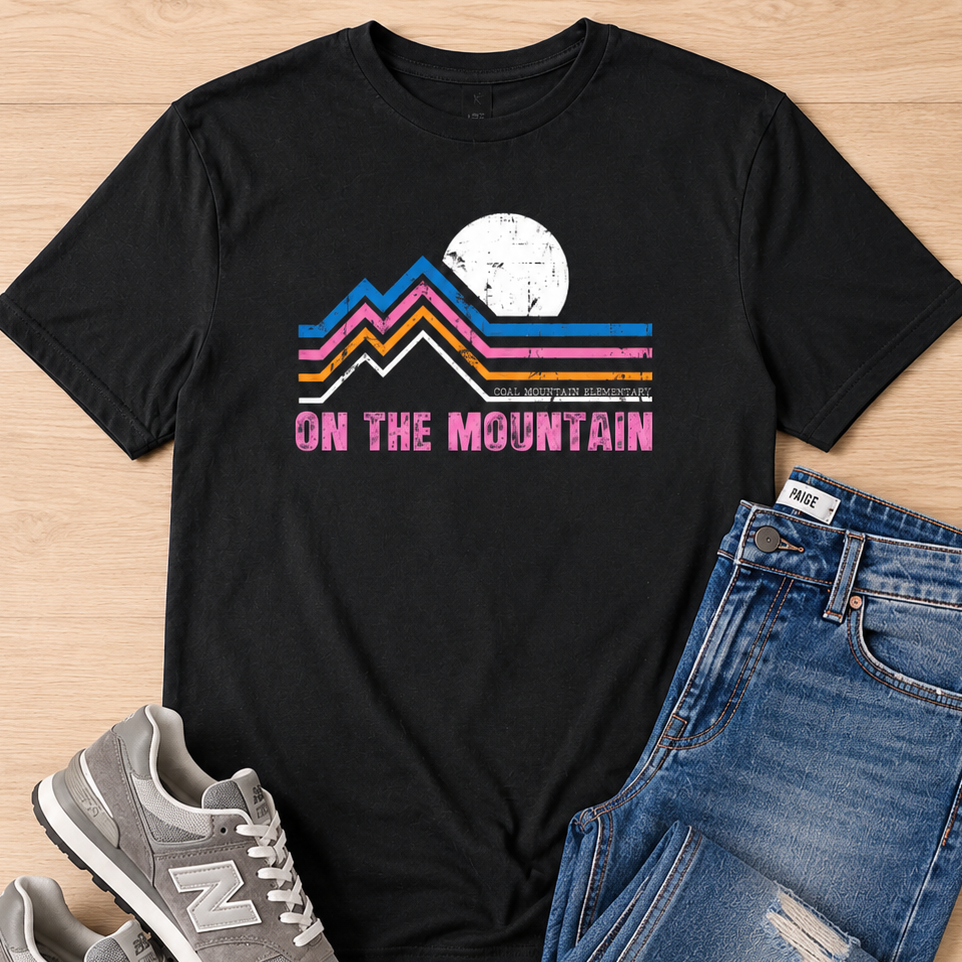 A black graphic t-shirt with a retro mountain design and the text 'ON THE MOUNTAIN' in pink, styled in a flat lay with blue jeans and gray New Balance sneakers on a light wood background.