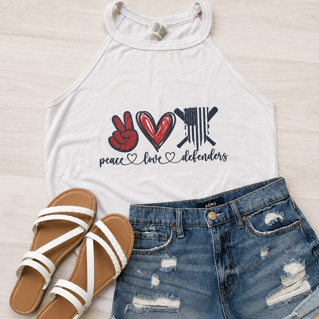 A white halter-style graphic tank top with a red and black 'peace love defenders' design, styled in a flat lay with distressed blue denim shorts and white strappy sandals on a light wood background.