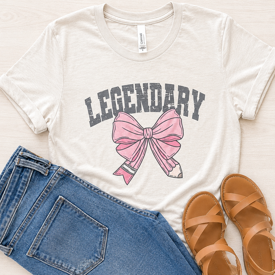 An off-white graphic t-shirt featuring the word 'LEGENDARY' in dark gray varsity letters above a pink bow illustration with a pencil tip. The t-shirt is styled in a flat lay with blue jeans, brown leather sandals, and a light wood background.