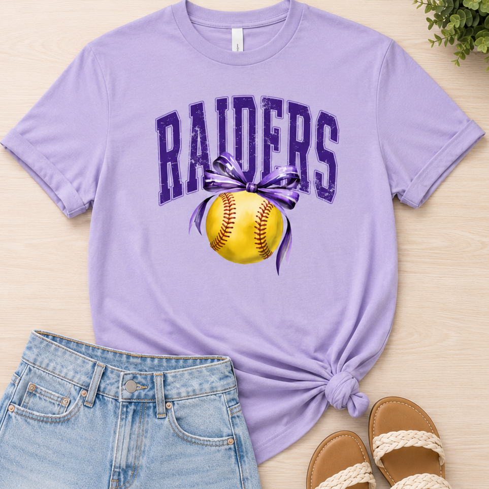 A flat-lay outfit featuring a light purple t-shirt with a 'RAIDERS' and softball graphic, styled with light blue denim shorts, brown sandals, and a small green plant on a light wood background.