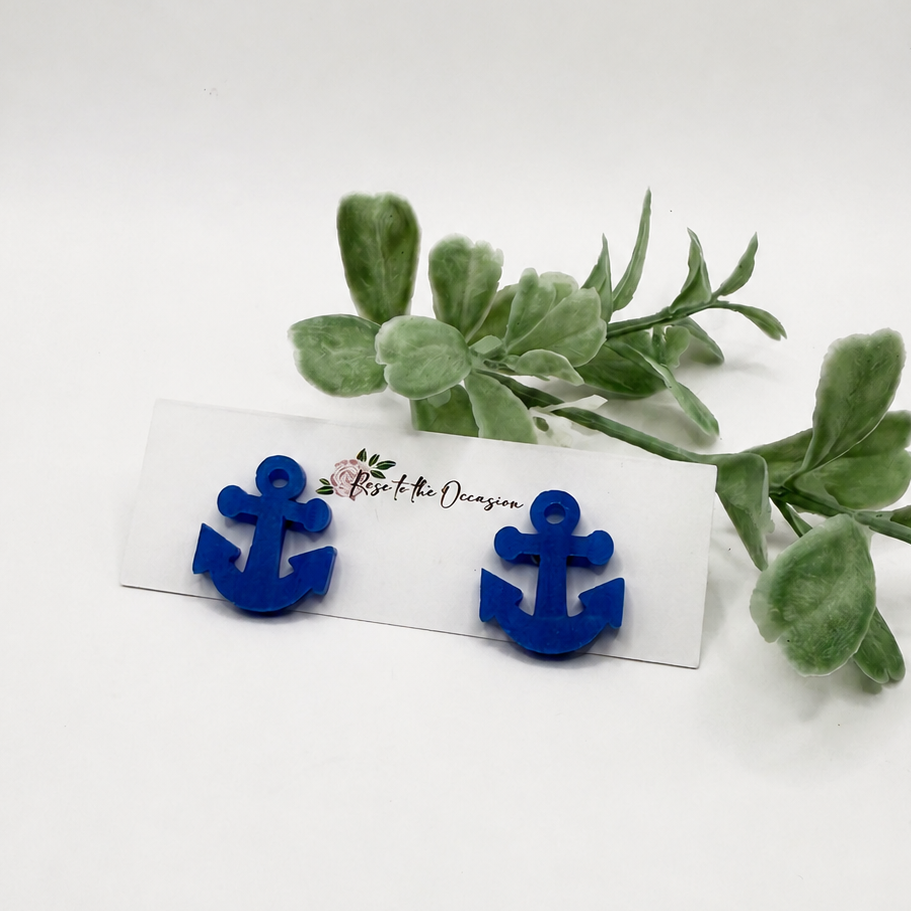 A pair of bright blue anchor-shaped stud earrings displayed on a small white card with a decorative script logo, styled with green leaves on a plain white background.