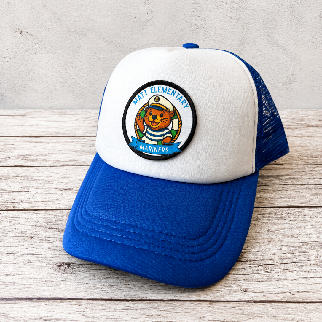 A blue and white trucker hat with a circular 'Hart Elementary Mariners' patch on the front, displayed on a rustic wooden surface against a textured gray wall.