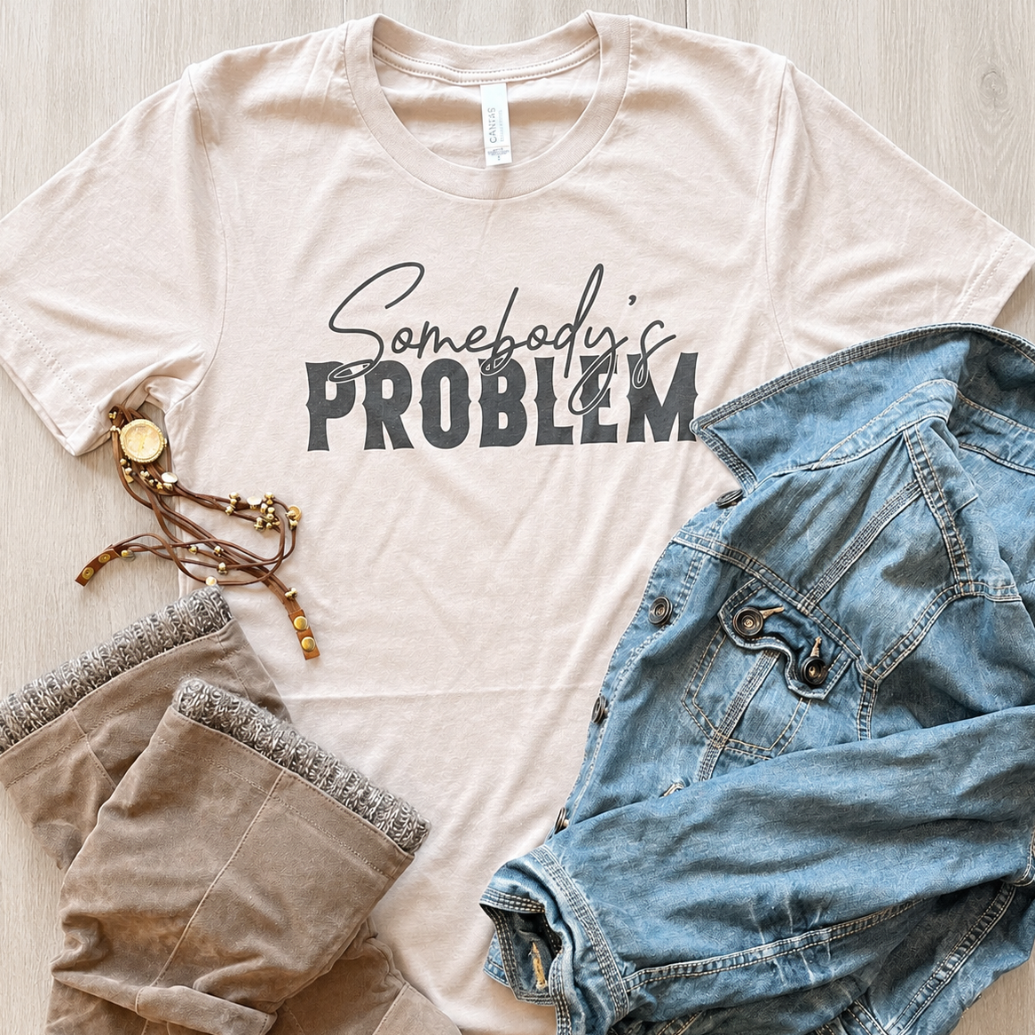 A beige graphic T-shirt laid flat on a light wood surface, styled with a blue denim jacket, tan pants, and a bracelet. The shirt has the phrase 'Somebody's PROBLEM' printed across the chest in black script and block lettering.