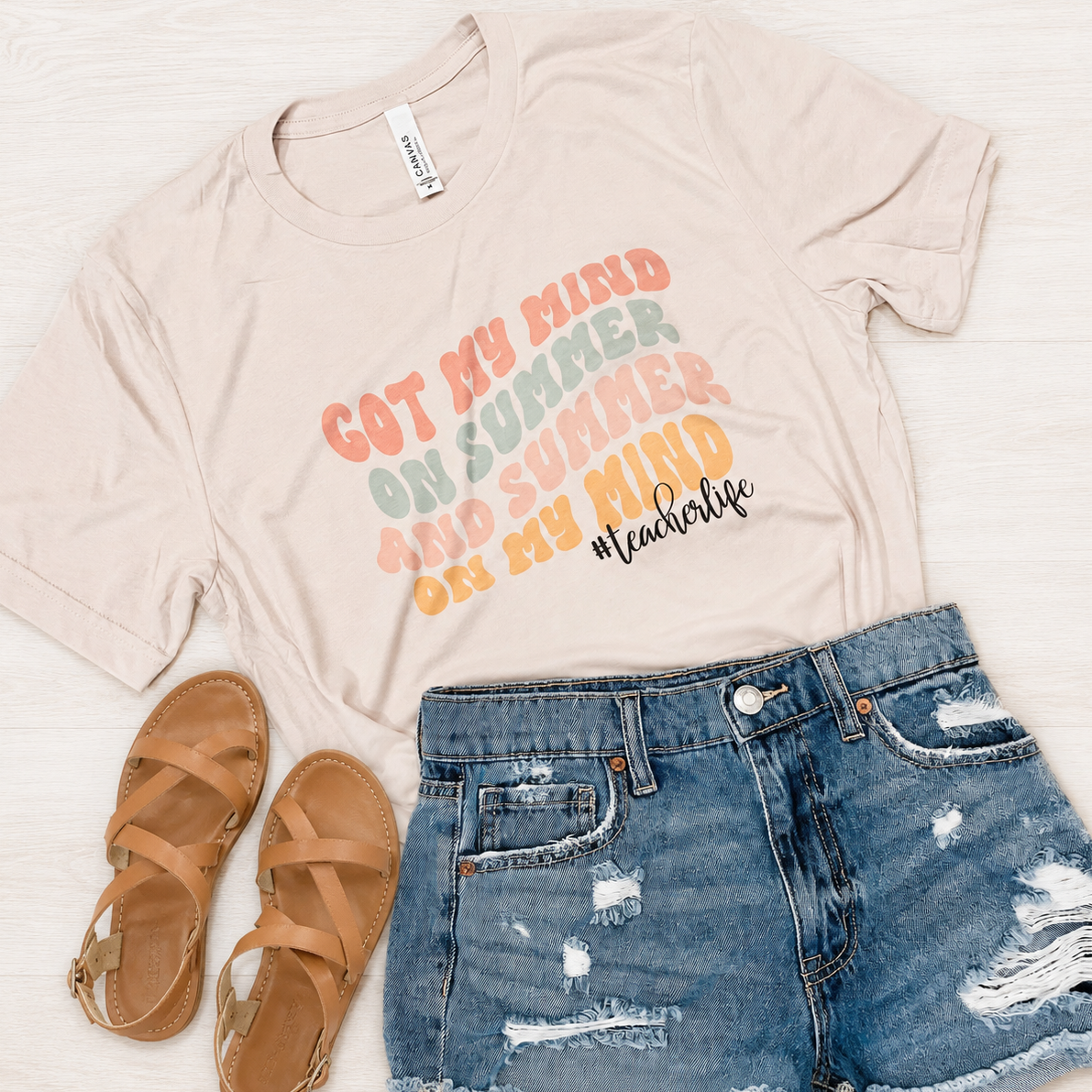 A styled flat-lay featuring a beige graphic t-shirt with the text 'GOT MY MIND ON SUMMER AND SUMMER ON MY MIND #teacherlife' in pastel colors, paired with distressed blue denim shorts and tan sandals on a white wood background.