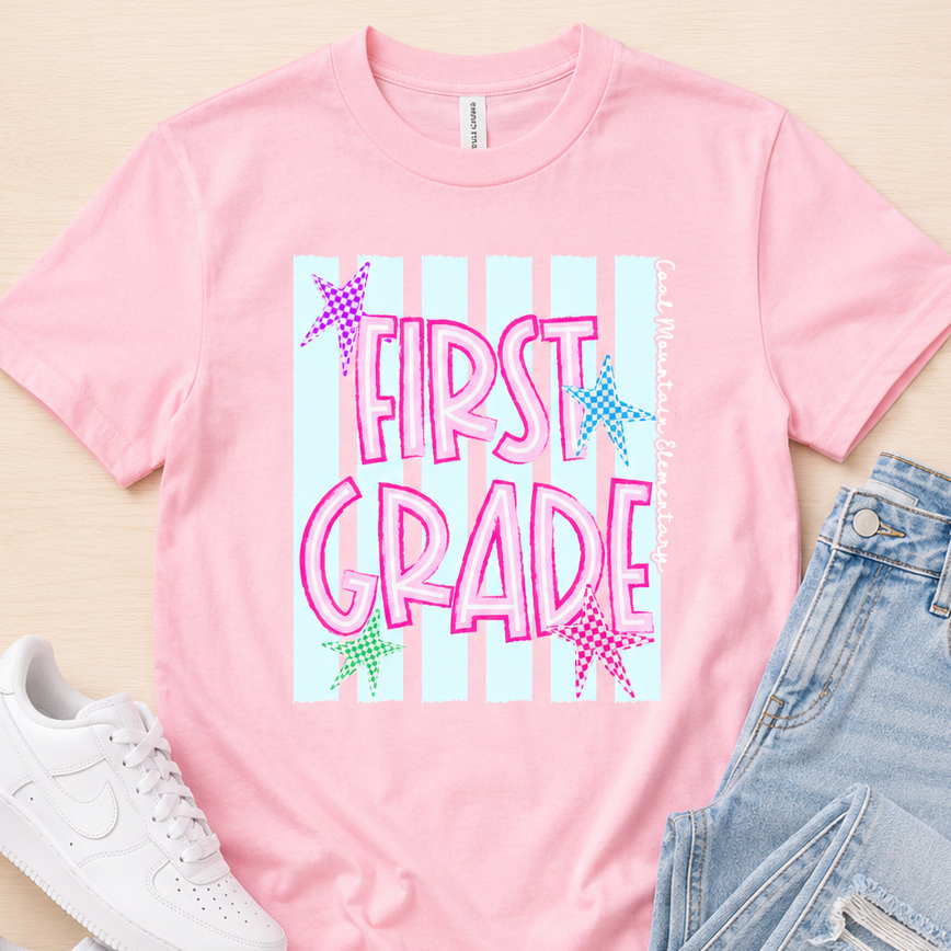 A pink graphic t-shirt with 'FIRST GRADE' in pink letters over a light blue and white striped background, styled in a flat lay with light blue jeans and white sneakers on a light wood background.