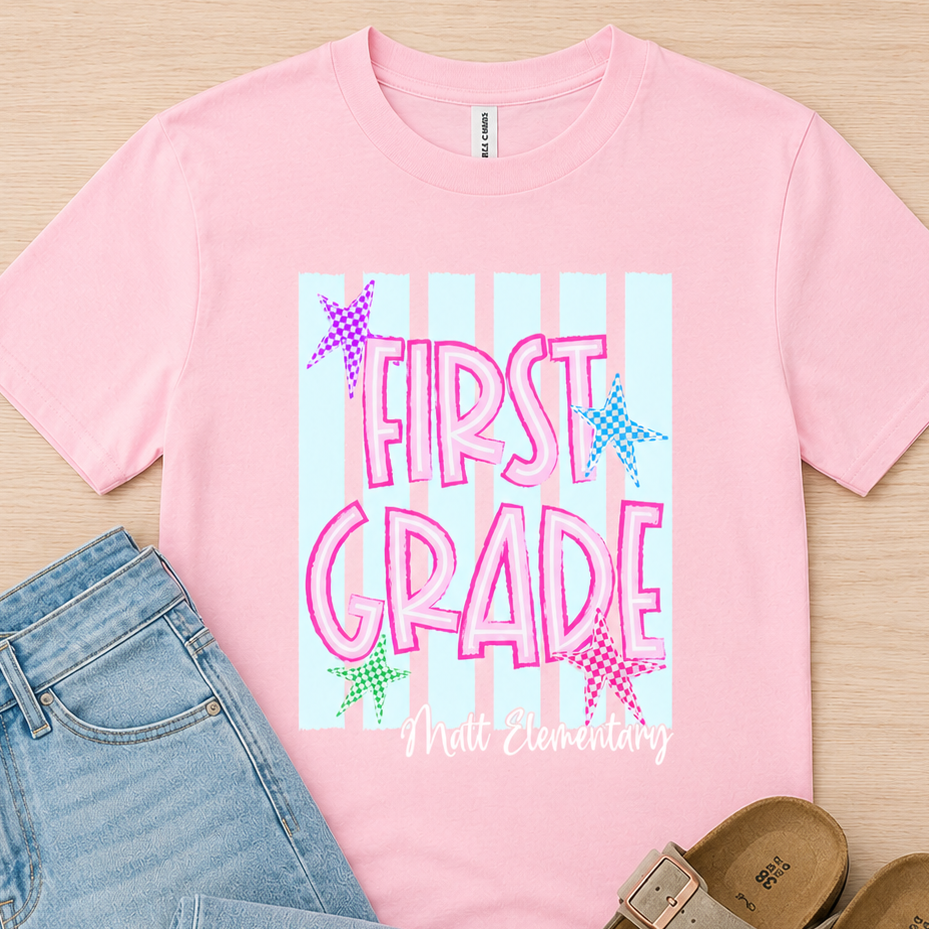 A pink graphic T-shirt laid flat on a light wood background, styled with light blue jeans and taupe slide sandals. The shirt has a large front graphic reading “FIRST GRADE” in pink lettering over pale blue vertical stripes with small multicolored star accents.