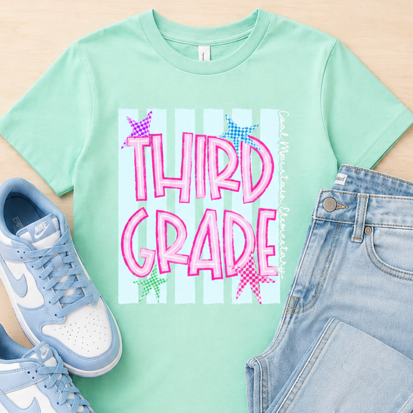 A mint green graphic t-shirt featuring the words 'THIRD GRADE' in pink bubble letters over a striped background, styled in a flat lay with light blue sneakers and light wash jeans on a light wood background.