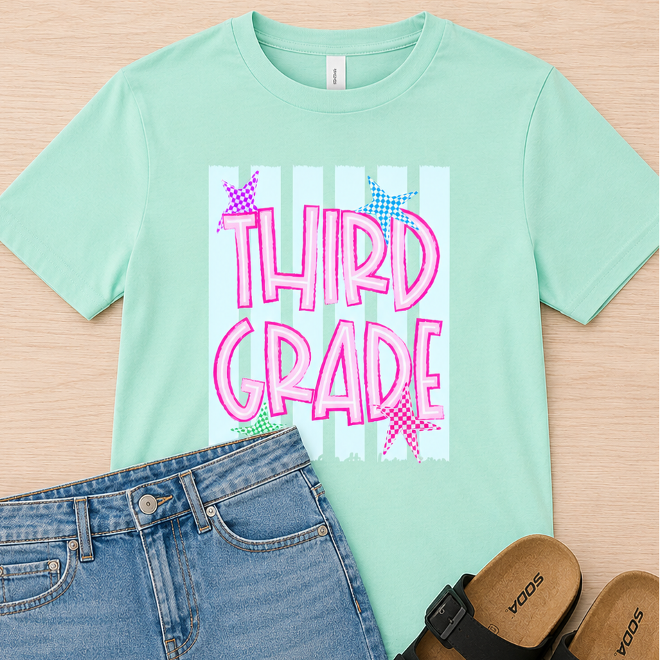 A mint green graphic t-shirt with 'THIRD GRADE' in pink bubble letters, styled in a flat lay with a blue denim skirt and black slide sandals on a light wood background.