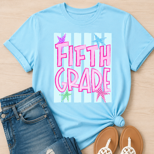 A light blue graphic t-shirt styled in a flat lay with distressed blue jeans and white sandals on a light wood background. The t-shirt features a 'FIFTH GRADE' graphic with pink lettering, white stripes, and colorful star accents.