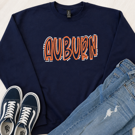 A navy blue crewneck sweatshirt with 'AU' printed across the chest in orange and white, styled in a flat lay with blue jeans and navy Vans sneakers on a light wood background.