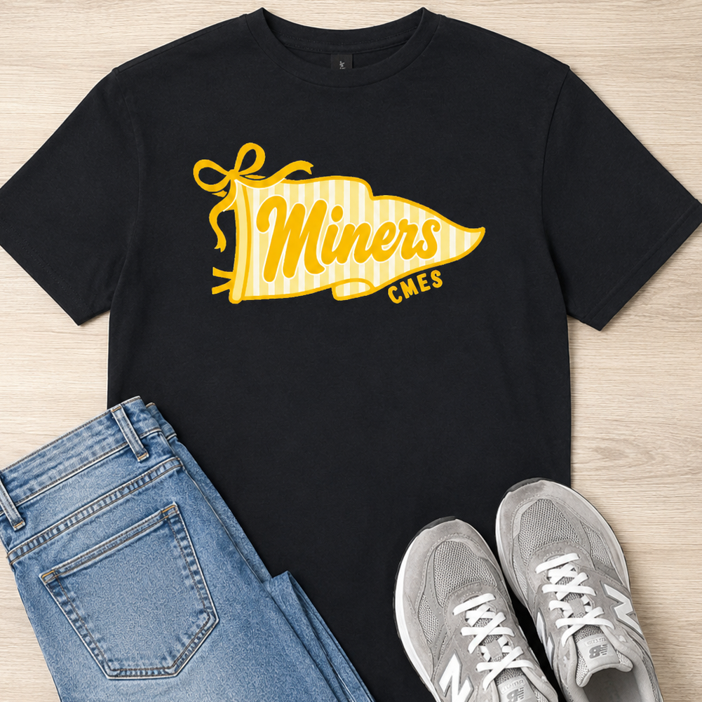 A black short-sleeve graphic T-shirt laid flat on a light wood background, styled with folded blue jeans and gray sneakers. The shirt has a large yellow and white pennant-style graphic reading "Miners" with smaller text "CMES" underneath.