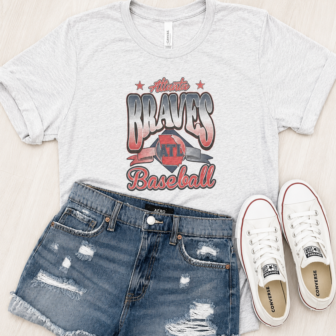 A light heather gray graphic T-shirt with distressed denim shorts and white Converse sneakers arranged on a light wood background. 