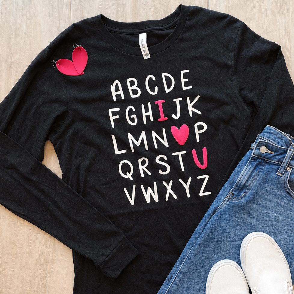 A black long-sleeve graphic T-shirt laid flat on a light wood surface, styled with blue jeans and white slip-on shoes. The shirt has a pink broken-heart graphic near the left chest and white alphabet letters with pink heart accents replacing some letters in the center.