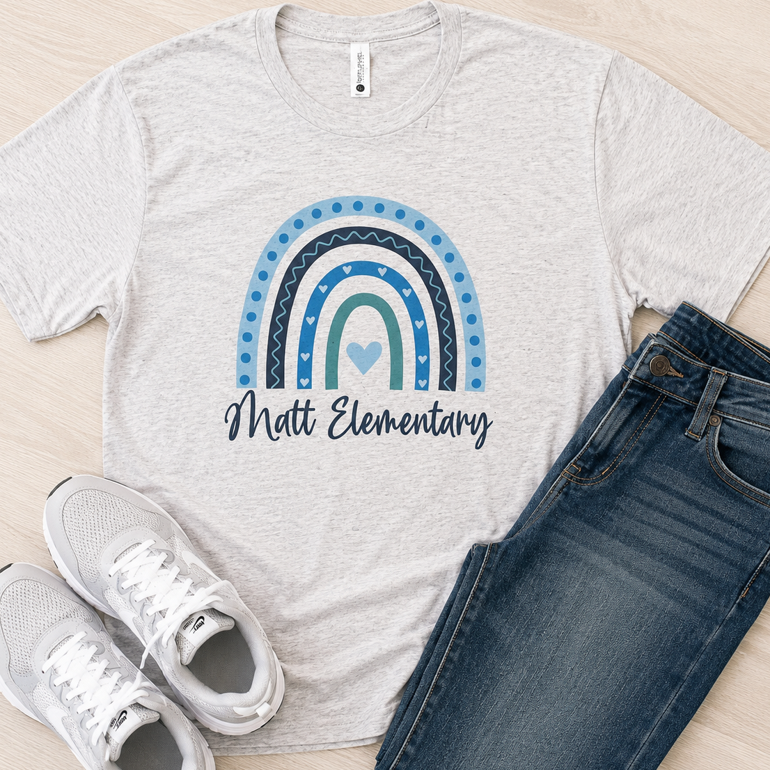 A light heather gray t-shirt with a blue and teal rainbow graphic and the text 'Matt Elementary' printed on the front, styled in a flat lay with blue jeans and white sneakers on a light wood floor.