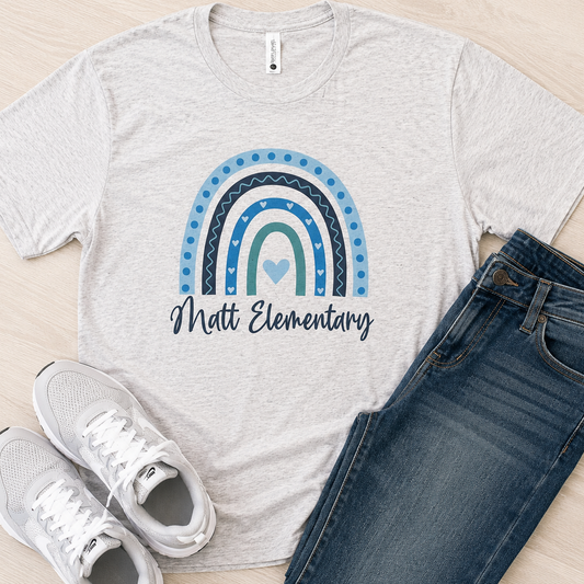 A light heather gray t-shirt with a blue and teal rainbow graphic and the text 'Matt Elementary' printed on the front, styled in a flat lay with blue jeans and white sneakers on a light wood floor.