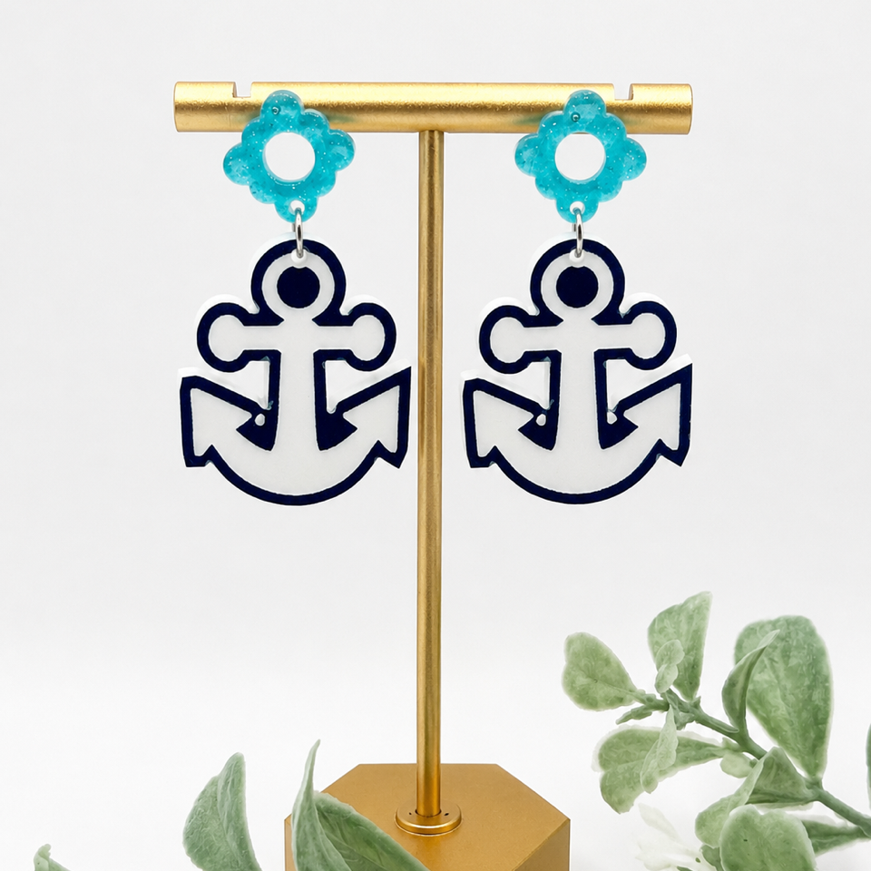 A pair of dangling earrings displayed on a gold stand against a white background, with decorative green leaves at the bottom. The earrings have turquoise flower-shaped studs and white anchor-shaped drops outlined in dark navy.