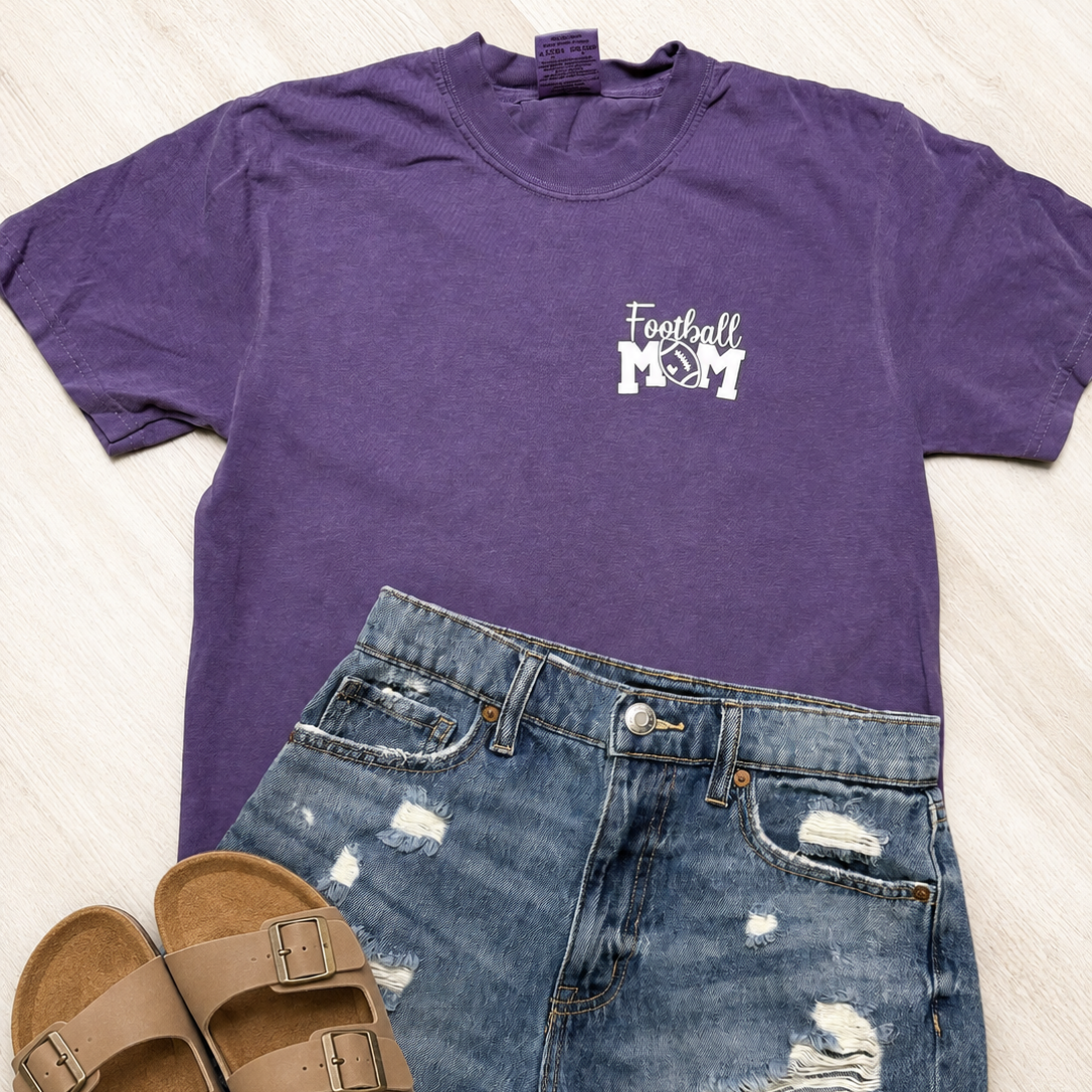 A purple short-sleeve t-shirt with a white 'Football Mom' graphic on the left chest, styled in a flat lay with distressed blue denim shorts and tan double-strap sandals on a light wood floor.