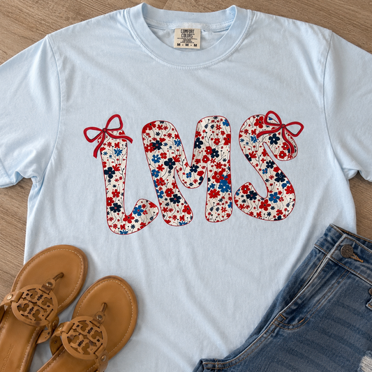 A light blue graphic T-shirt laid flat on a wood surface, styled with tan sandals and blue jeans. The shirt has a large floral 'LMS' graphic with red bows on the letters.