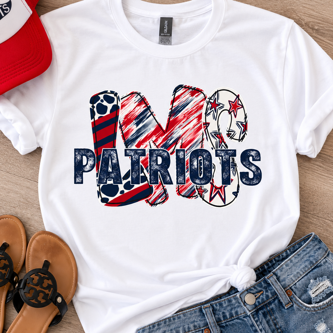 White short-sleeve graphic T-shirt laid flat on a wood surface, styled with a red-and-white cap, tan sandals, and blue jeans. The shirt has a large 'MO PATRIOTS' text graphic in red, white, and blue with sports-themed lettering details.