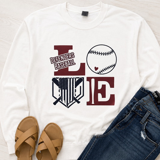 A white long-sleeve sweatshirt laid flat on a light wood surface, styled with tan sandals and blue jeans. The sweatshirt has a large graphic spelling LOVE, where the O is a baseball and the letters include a 'Defenders Baseball' shield logo.