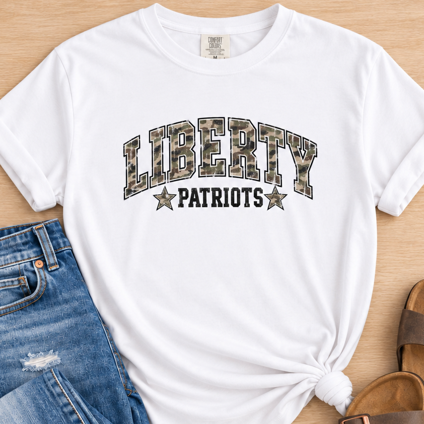 A white short-sleeve T-shirt with 'LIBERTY PATRIOTS' printed across the front in camouflage and black lettering, styled in a flat lay with blue jeans and brown slide sandals on a light wood background.