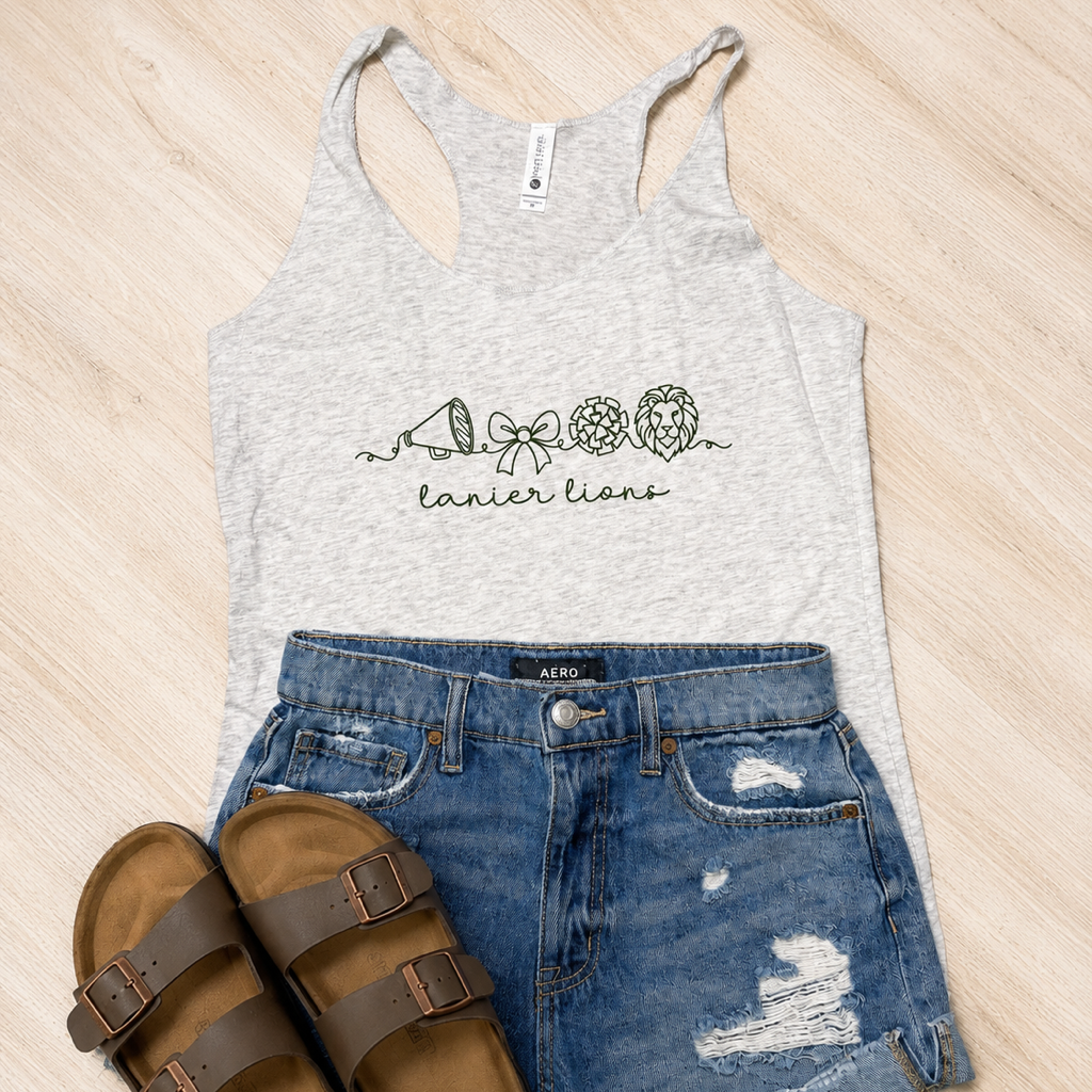 A styled flat-lay of a women's heather gray racerback tank top with a black line art graphic reading 'tanier lions' and 'Lions' with a lion head. The tank top is paired with distressed blue denim shorts and brown slide sandals on a light wood background.