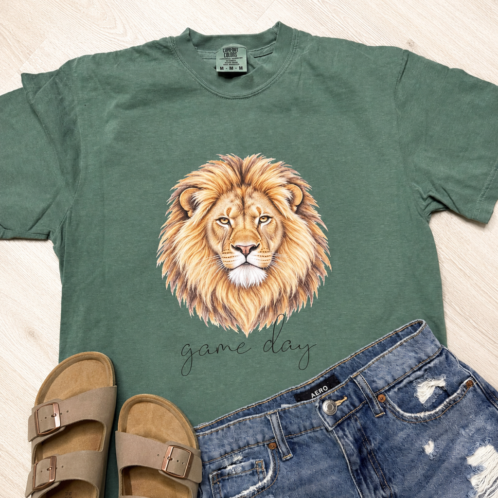A styled flat-lay featuring a sage green t-shirt with a detailed lion graphic and the text 'game day', paired with blue distressed denim shorts and taupe double-strap sandals on a light wood floor background.