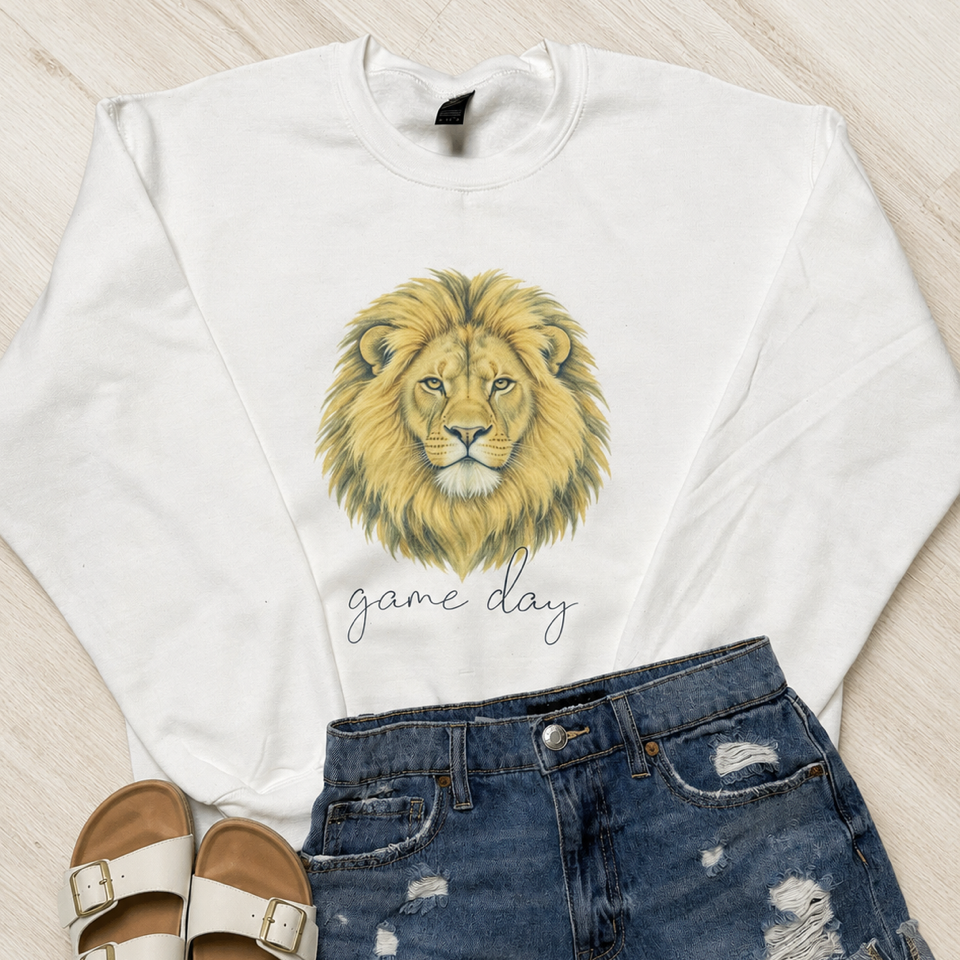 A white crewneck sweatshirt featuring a watercolor-style golden lion graphic and the text 'game day' in script, styled in a flat lay with distressed blue denim shorts and beige double-strap sandals on a light wood floor.